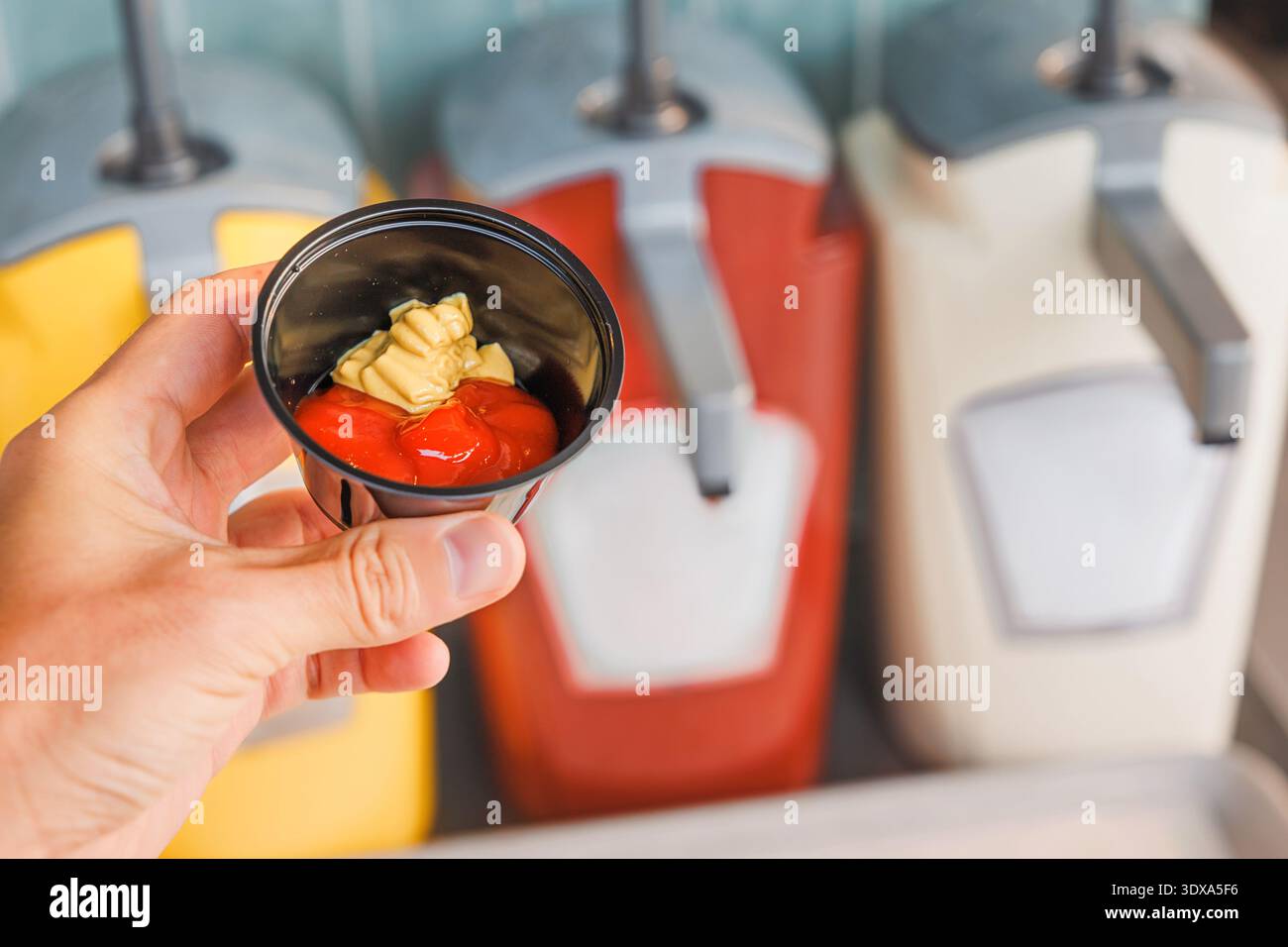 Hand holding a small plastic cup filled with red ketchup and yellow mustard, with condiment dispensers in the background Stock Photo