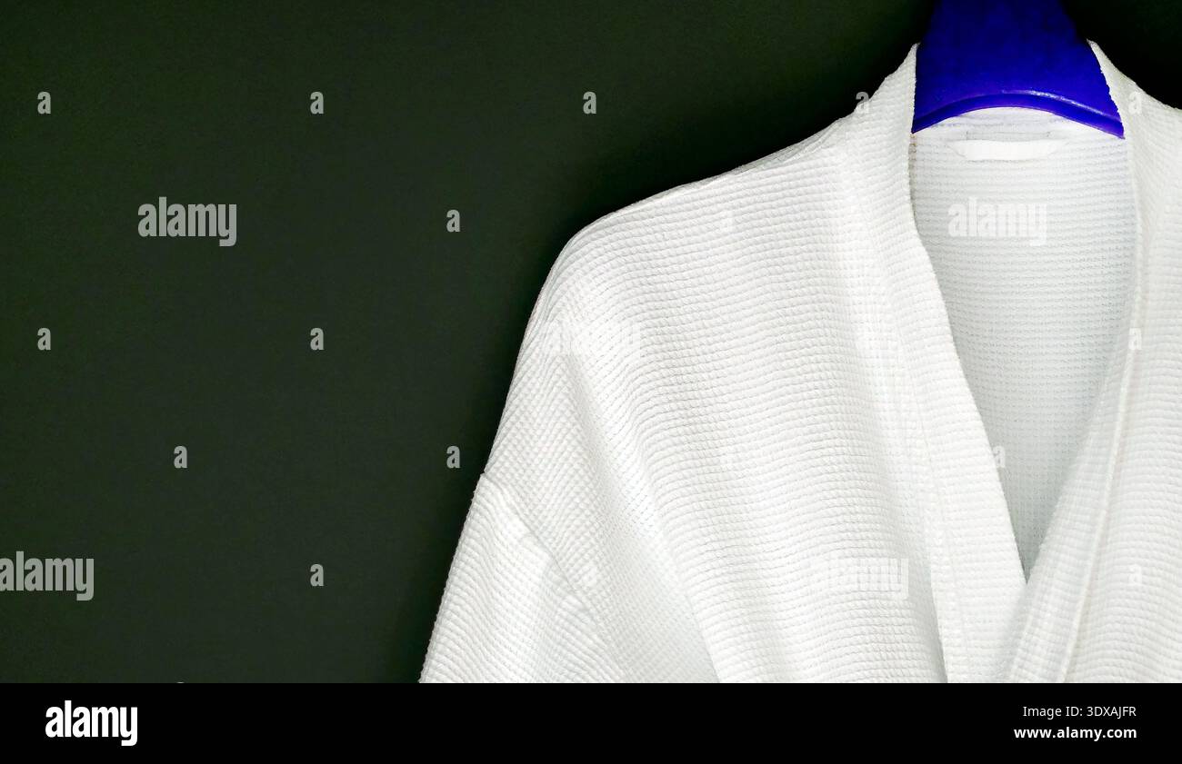 Clean white cotton bathrobe with waffle weave texture hanging on a blue hanger against a black wall. Minimalist composition with ample negative space Stock Photo