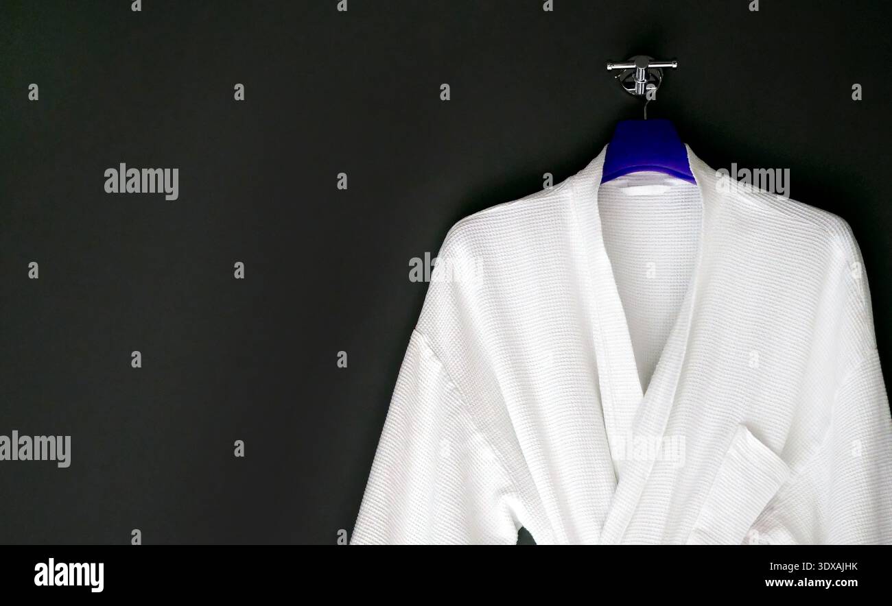 Clean white cotton bathrobe with waffle weave texture hanging on a blue hanger against a black wall. Minimalist composition with ample negative space Stock Photo