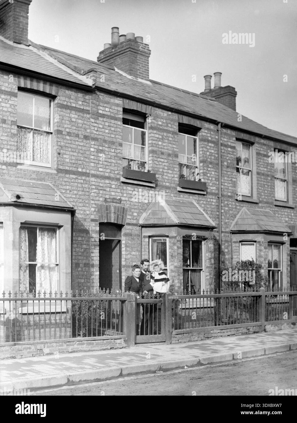 A vintage photograph of a family posing in front of their brick terraced home, featuring period architectural details like bay windows, iron railings, and lace curtains. Stock Photo