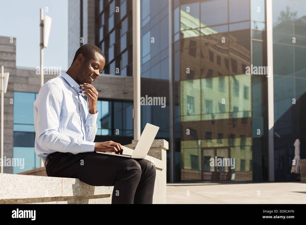 Young Man Works on Laptop Outdoors in a Modern City Setting by a Glass Building Stock Photo