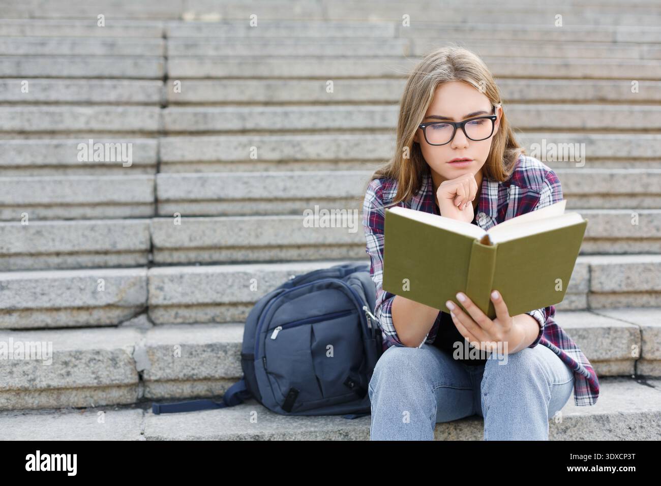 Student Reads a Book While Sitting on Stone Steps With a Backpack Beside Her Stock Photo