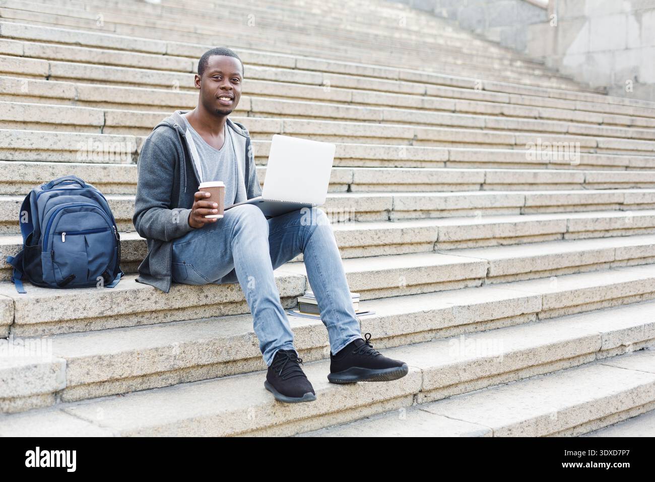 Young Man Sits on Stairs Using Laptop and Drinking Coffee in Urban Setting During Daytime Stock Photo