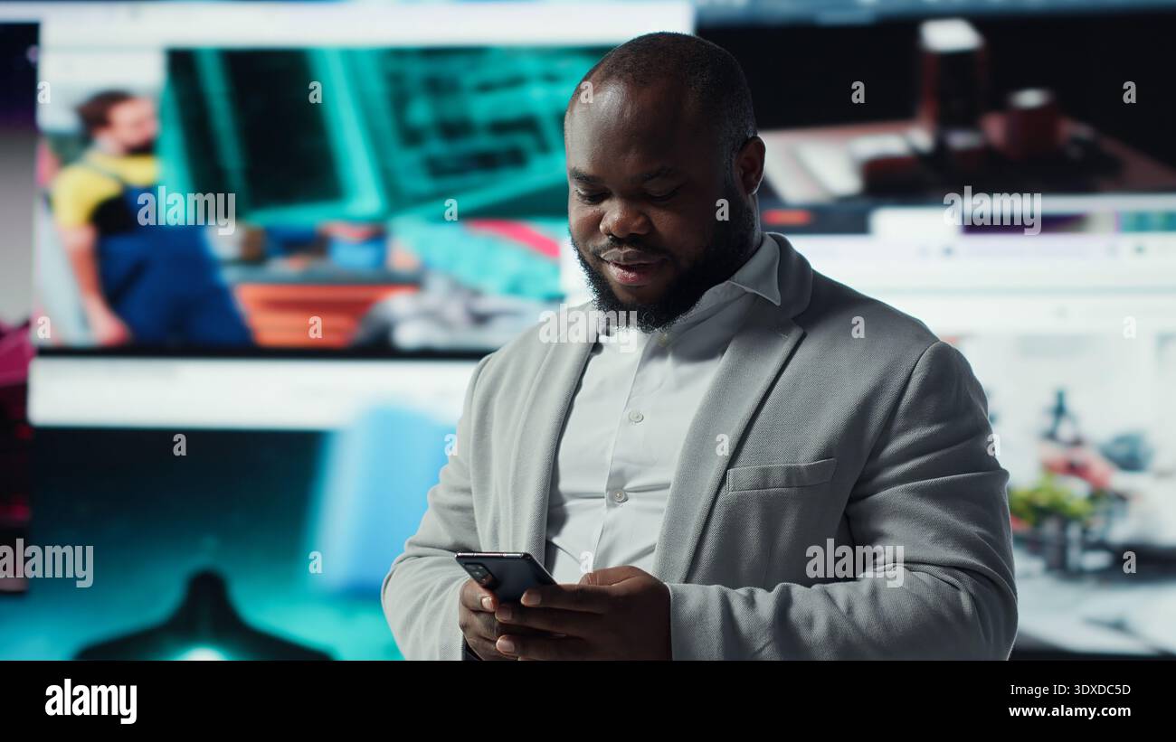 Portrait of cheerful man browsing social media feed on smartphone in front of digital video wall. Happy person using mobile phone, floating icons highlight interactive engagement online, camera A Stock Photo