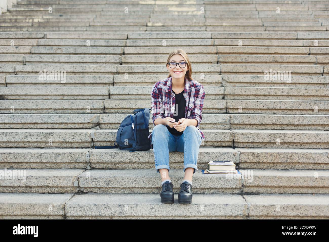 Young Person Sits on Steps With Books and Backpack While Using a Phone Stock Photo
