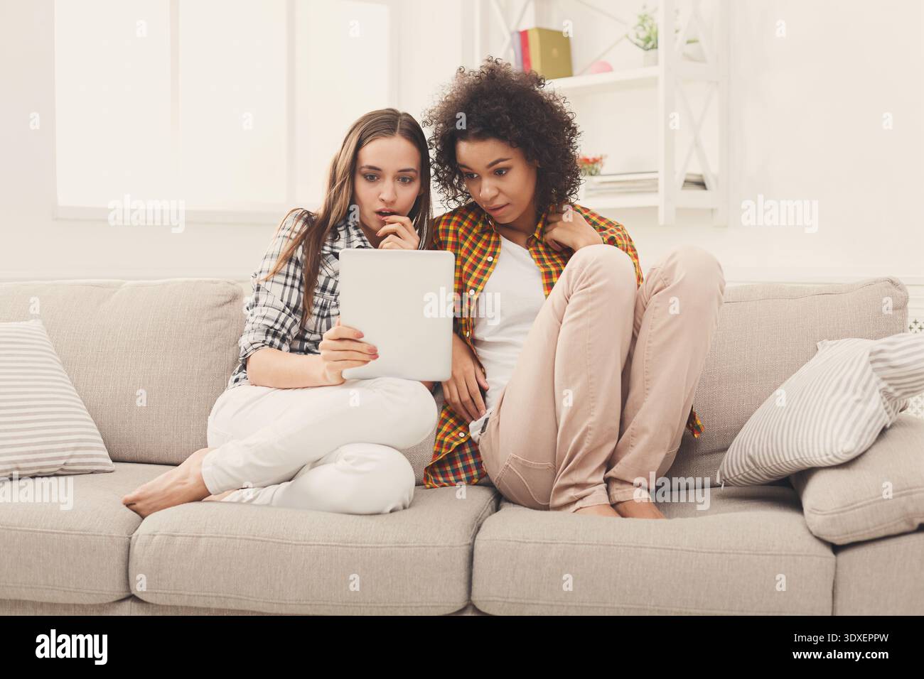 Two Friends Sit Together on a Couch While Looking at a Tablet and Sharing an Experience Stock Photo