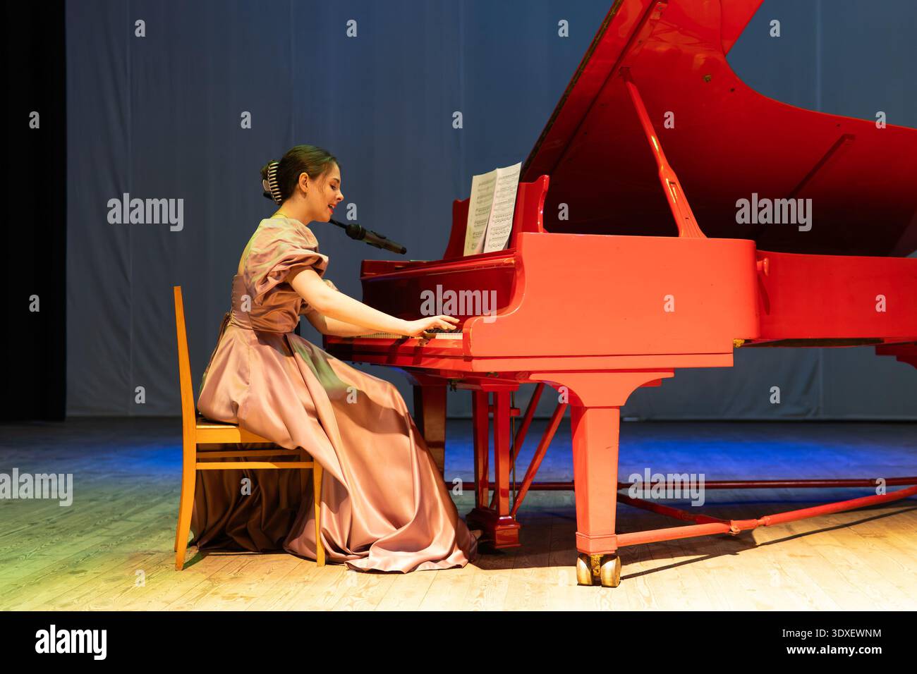 Young woman playing grand piano on stage. Elegant live performance, focused and expressive, capturing musical talent and stage presence. Stock Photo
