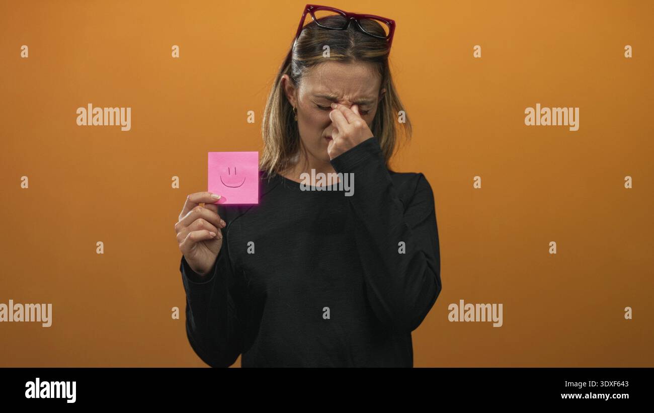 Young blonde woman pinches nose with thumb and forefinger while holding pink sticky note with smile drawing against orange studio wall, wearing black Stock Photo