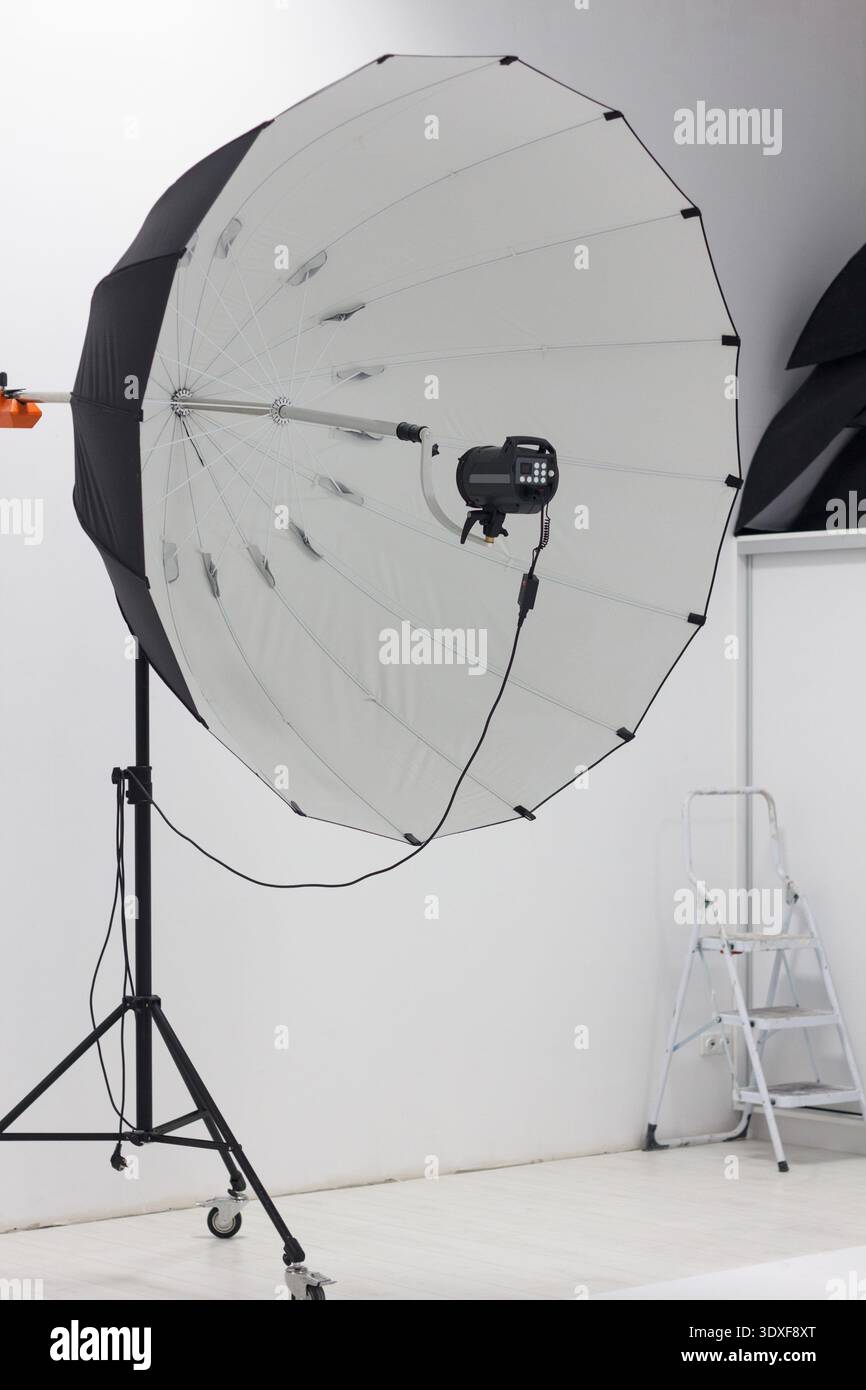 Professional photography studio setup. A large parabolic umbrella softbox with a strobe flash is on a stand in a minimalist white room. Behind the sce Stock Photo