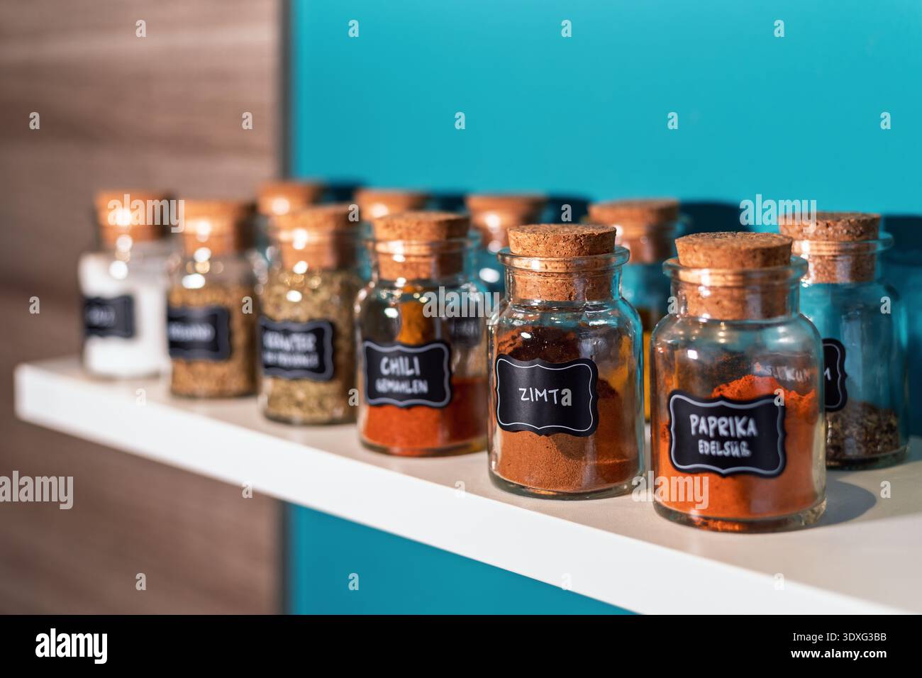 Glass spice jars with cork lids arranged on a white kitchen shelf. The jar labeled cinnamon in German is in focus. Stock Photo