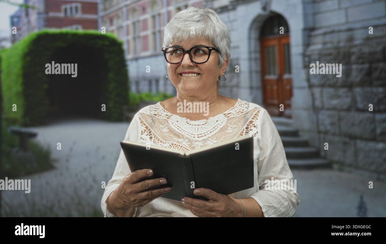 Woman with grey hair holding a black book, hands visible, reading by a stone building entrance in a courtyard; serenity reflection. Stock Photo
