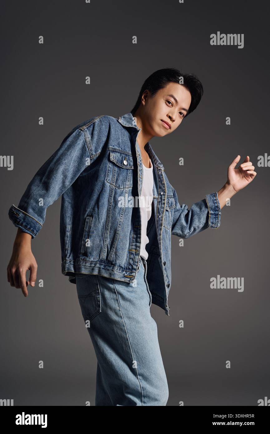 Young Asian man confidently poses in denim, showcasing style in a grey studio. Stock Photo