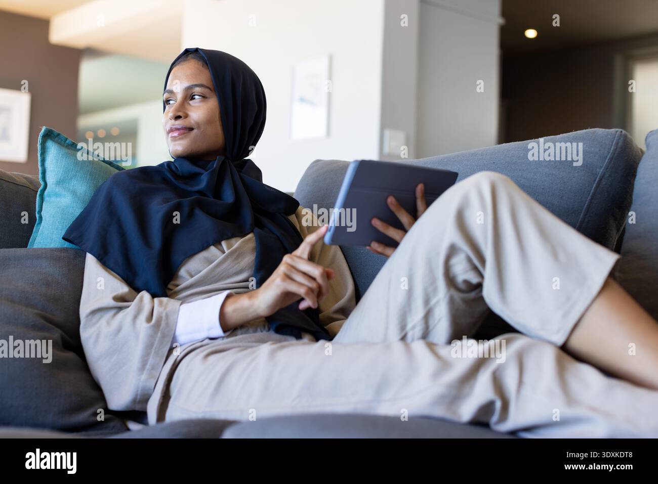Woman reclining on sofa with teal pillow holding dark tablet tapping screen wearing navy headscarf Stock Photo