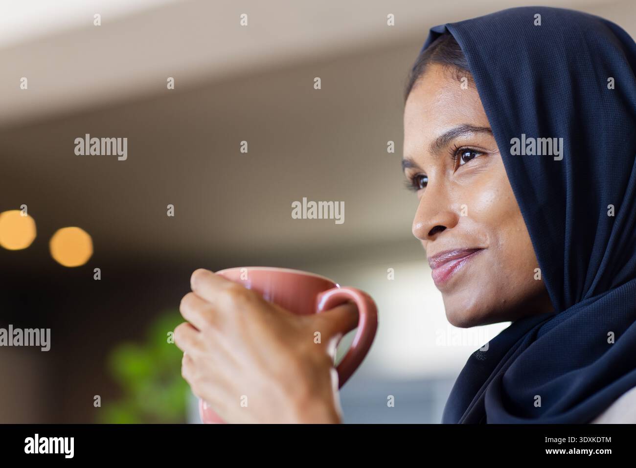 Woman wearing dark navy headscarf holding pink ceramic mug near mouth, smiling in cafe, copy space Stock Photo
