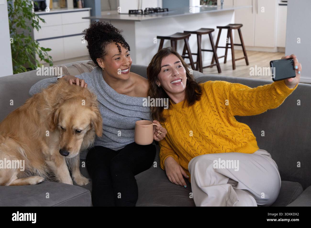 Diverse female friends on gray sectional couch at home taking phone selfie holding mug petting dog Stock Photo