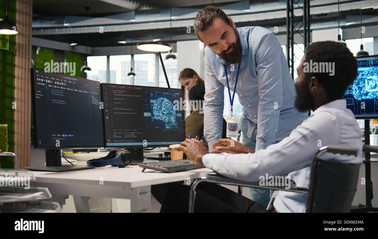 Manager supervising developer with paraplegia writing code, developing deep learning APIs. Supervisor overseeing work done by IT expert in wheelchair troubleshooting software errors using AI, camera A Stock Photo