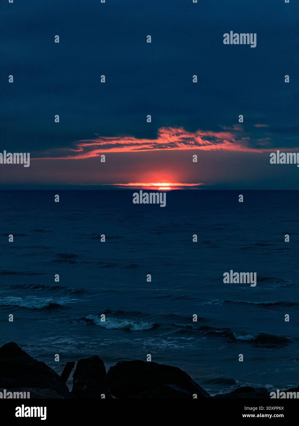 Dramatic winter sunrise over the English Channel from Worthing beach in West Sussex, fiery red sun rising above dark cloud bank across deep blue sea a - Smartphone Captured Stock Image