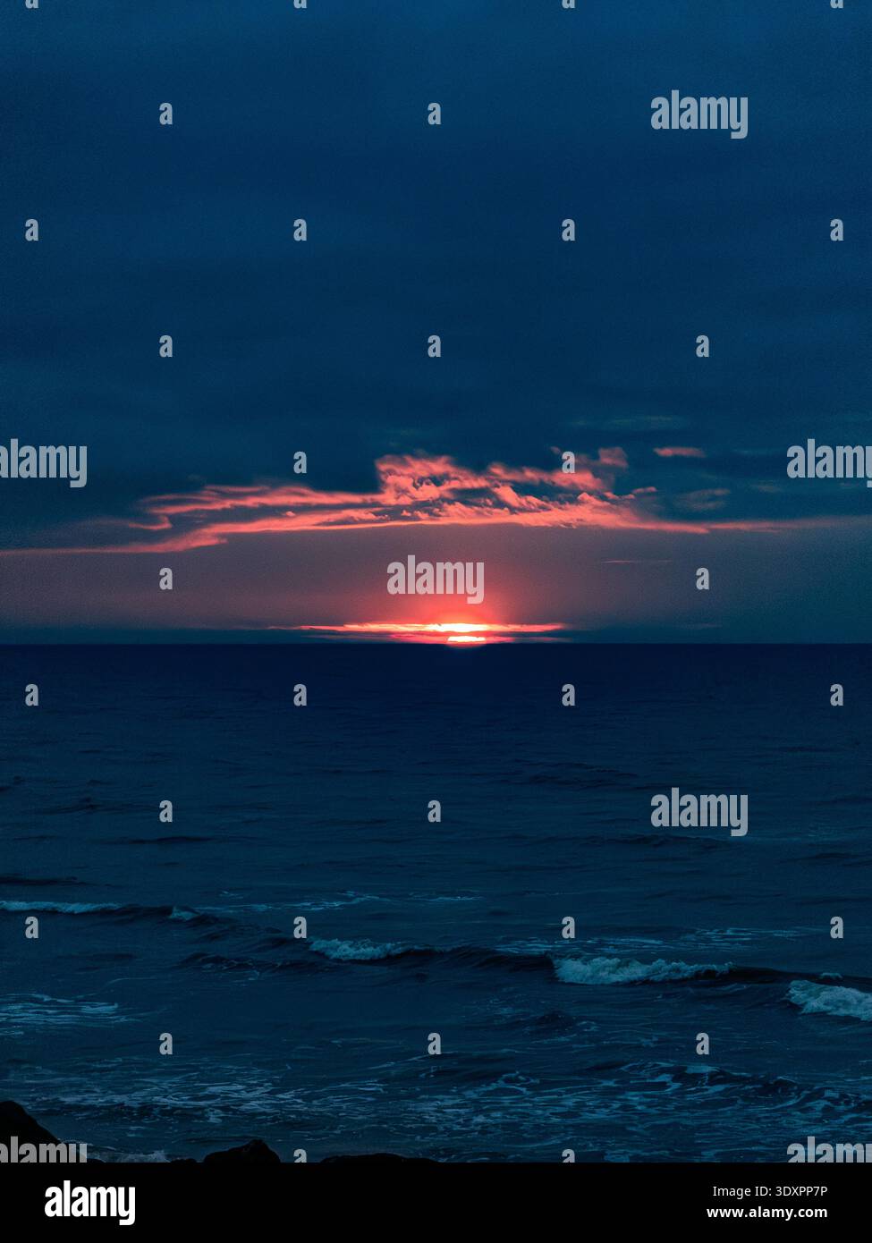 Dramatic winter sunrise over the English Channel from Worthing beach in West Sussex, fiery red sun rising above dark cloud bank across deep blue sea - Smartphone Captured Stock Image