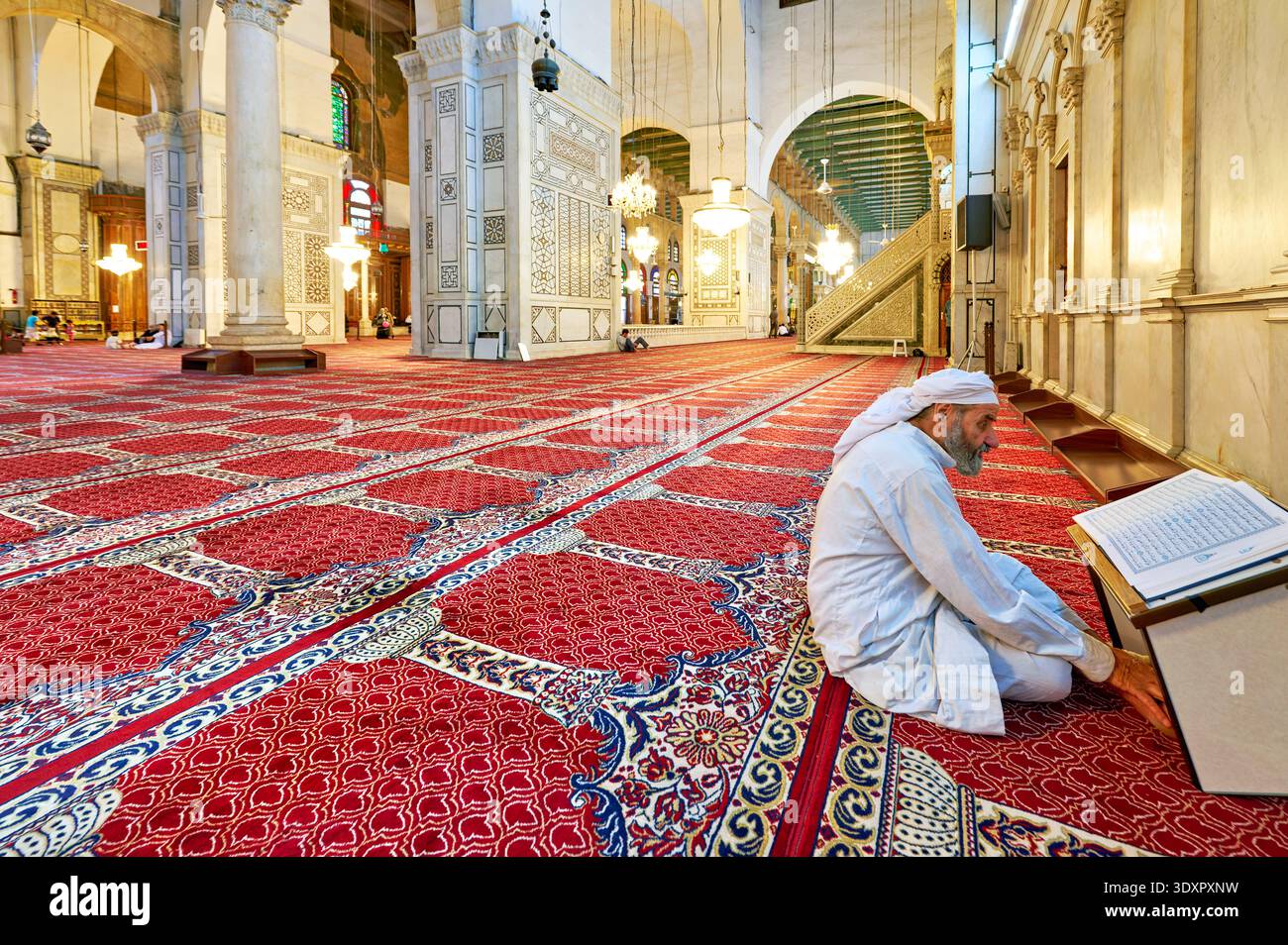 A man sits on the carpet inside Umayyad Mosque in Damascus, reading the Quran. The mosque has intricate designs and high ceilings. Stock Photo