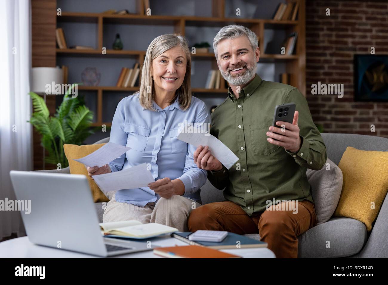 Happy senior couple sitting on a sofa, holding bills and a smartphone while using a laptop to review and manage expenses, representing financial planning and successful budgeting at home Stock Photo