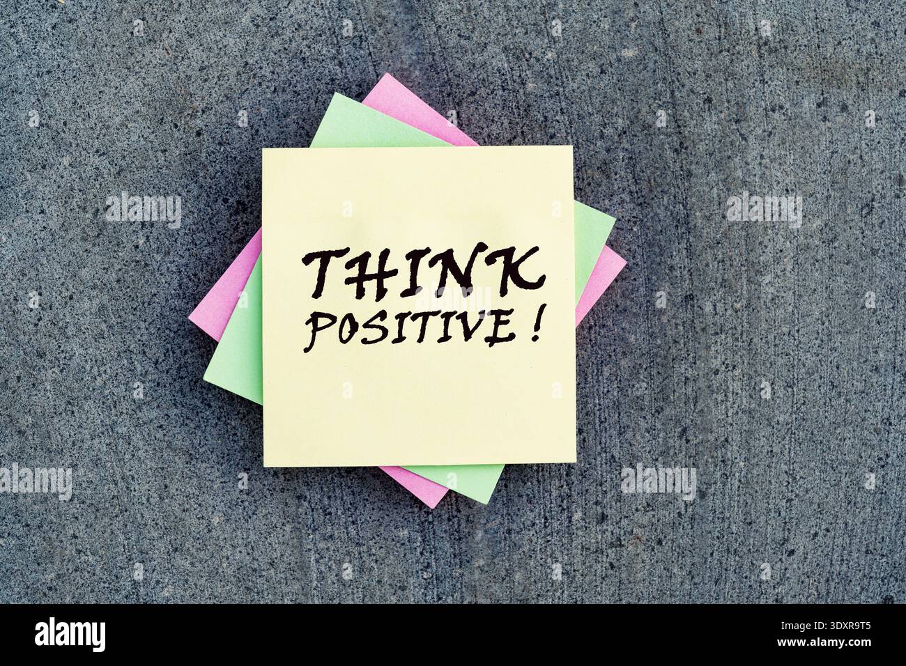 "THINK POSITIVE!" handwritten on a stack of colorful sticky notes over a grey stone background. Stock Photo