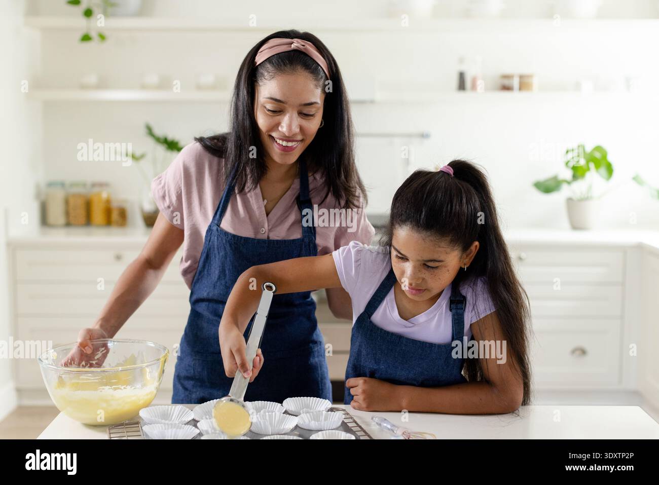 African American mom and child scooping batter from glass bowl into paper liners at home counter Stock Photo