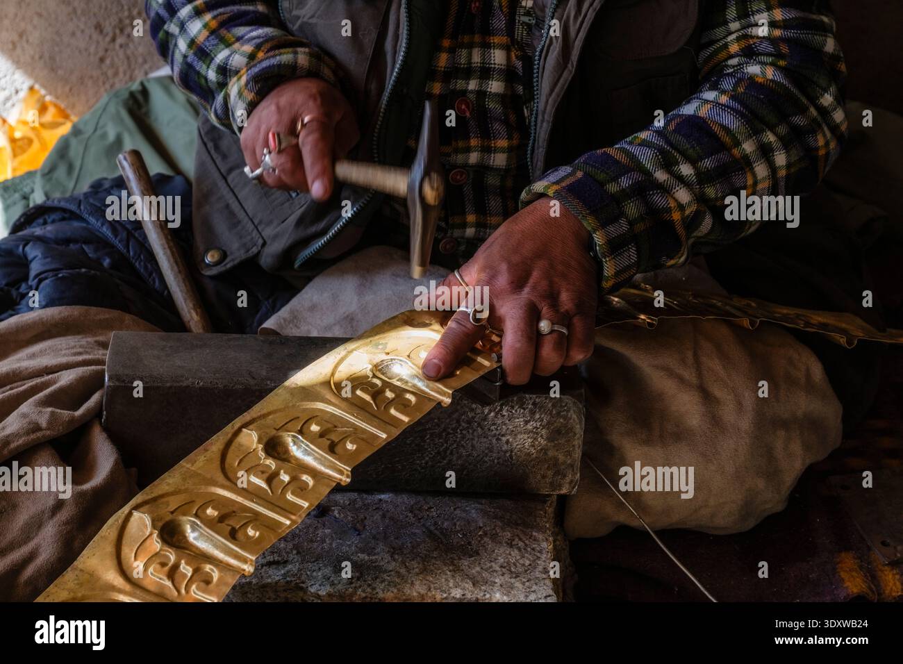 Close-up of a craftsman at Matho using a hammer to emboss traditional patterns into a long strip of golden metal on a stone anvil. Stock Photo