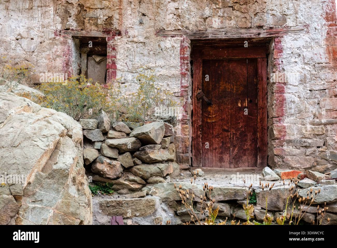 A weathered red wooden door and a small square window are set into the textured, whitewashed stone walls of Hemis Gompa in Hemis. Stock Photo