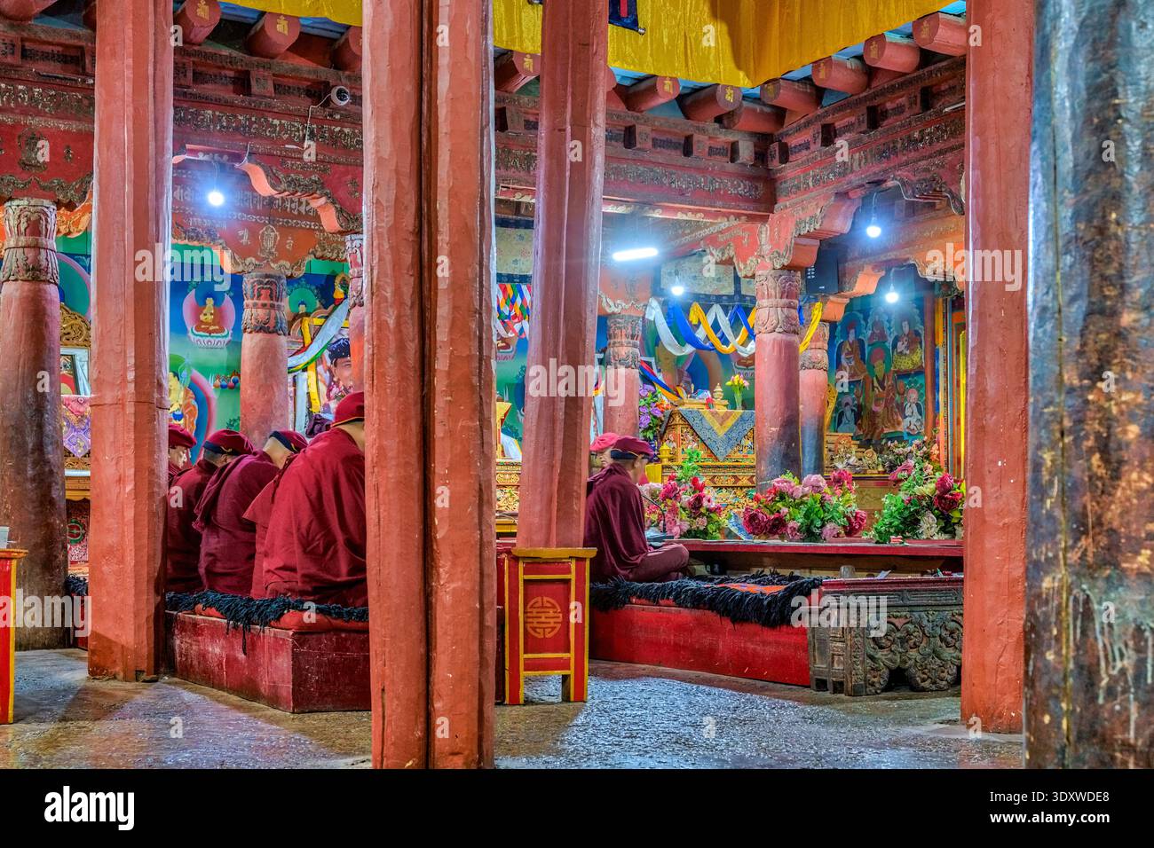Buddhist monks in red robes sit in rows between thick red pillars ...