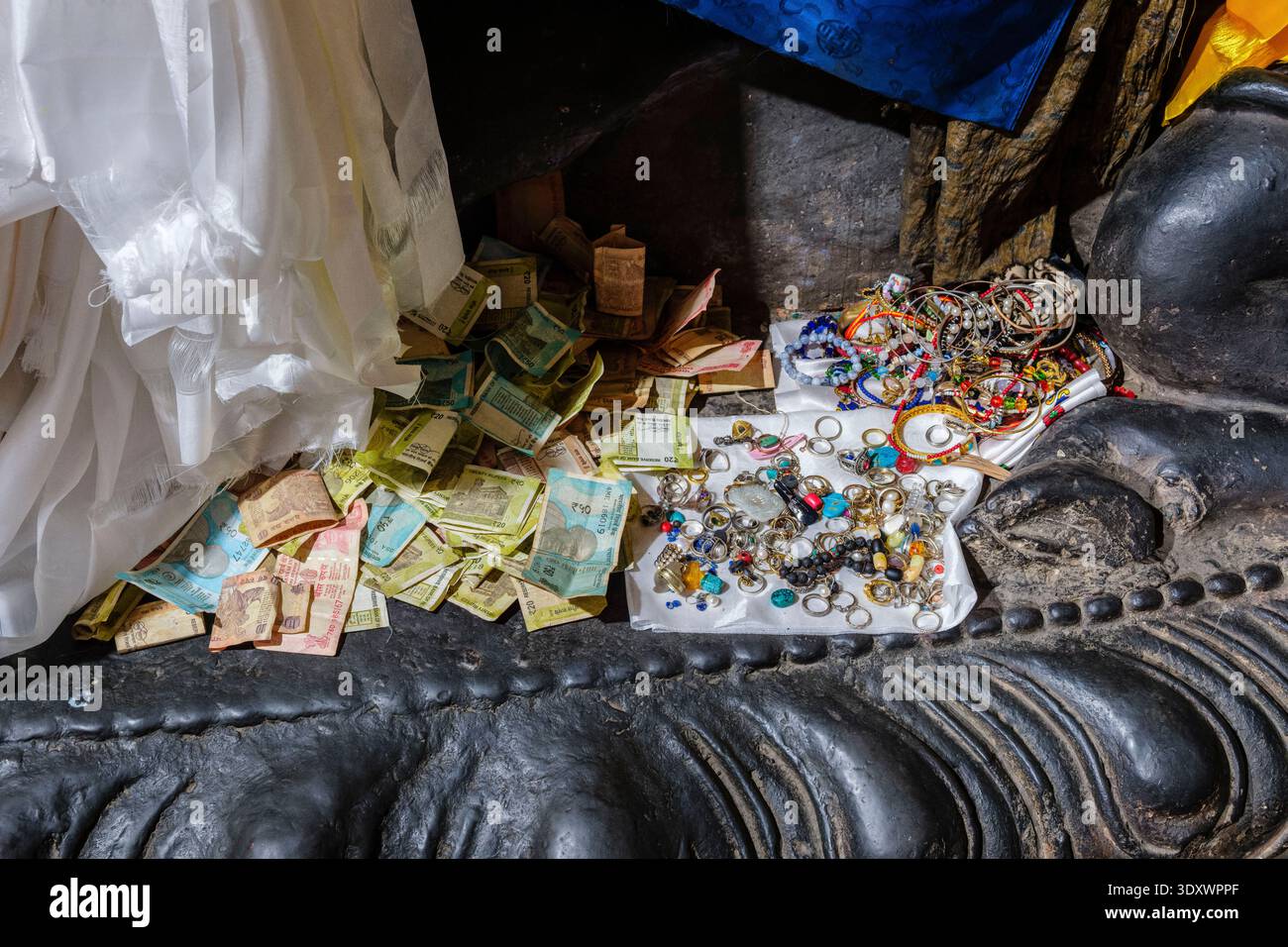 Offerings of Indian Rupee banknotes and various pieces of jewellery placed at the feet of a religious statue on a dark plinth in Thiksey Gompa. Stock Photo
