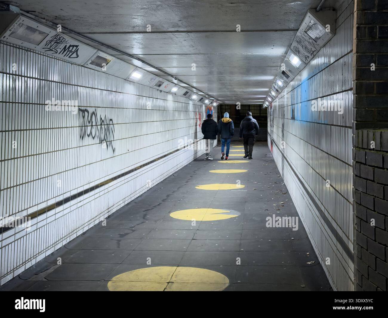 Three people walking away through a dimly lit underground pedestrian subway tunnel with tiled walls and bright circular floor markings coventry, uk - Smartphone Captured Stock Image