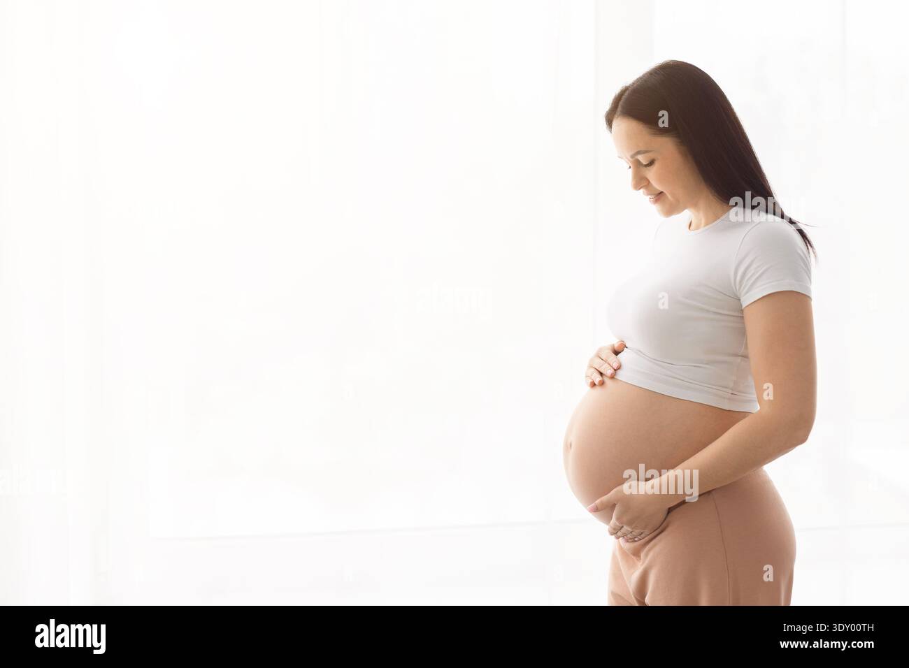 Side view of young pregnant woman holding belly in bright home interior Stock Photo
