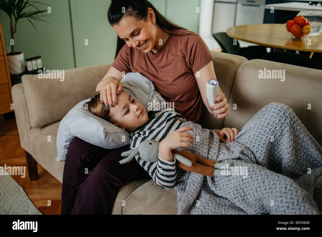 Caring mother measuring temperature of her ill son lying on sofa  Stock Photo