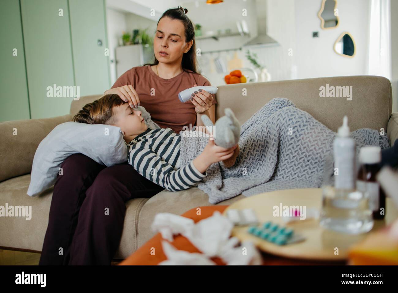 Caring mother measuring temperature of her ill son lying on sofa  Stock Photo