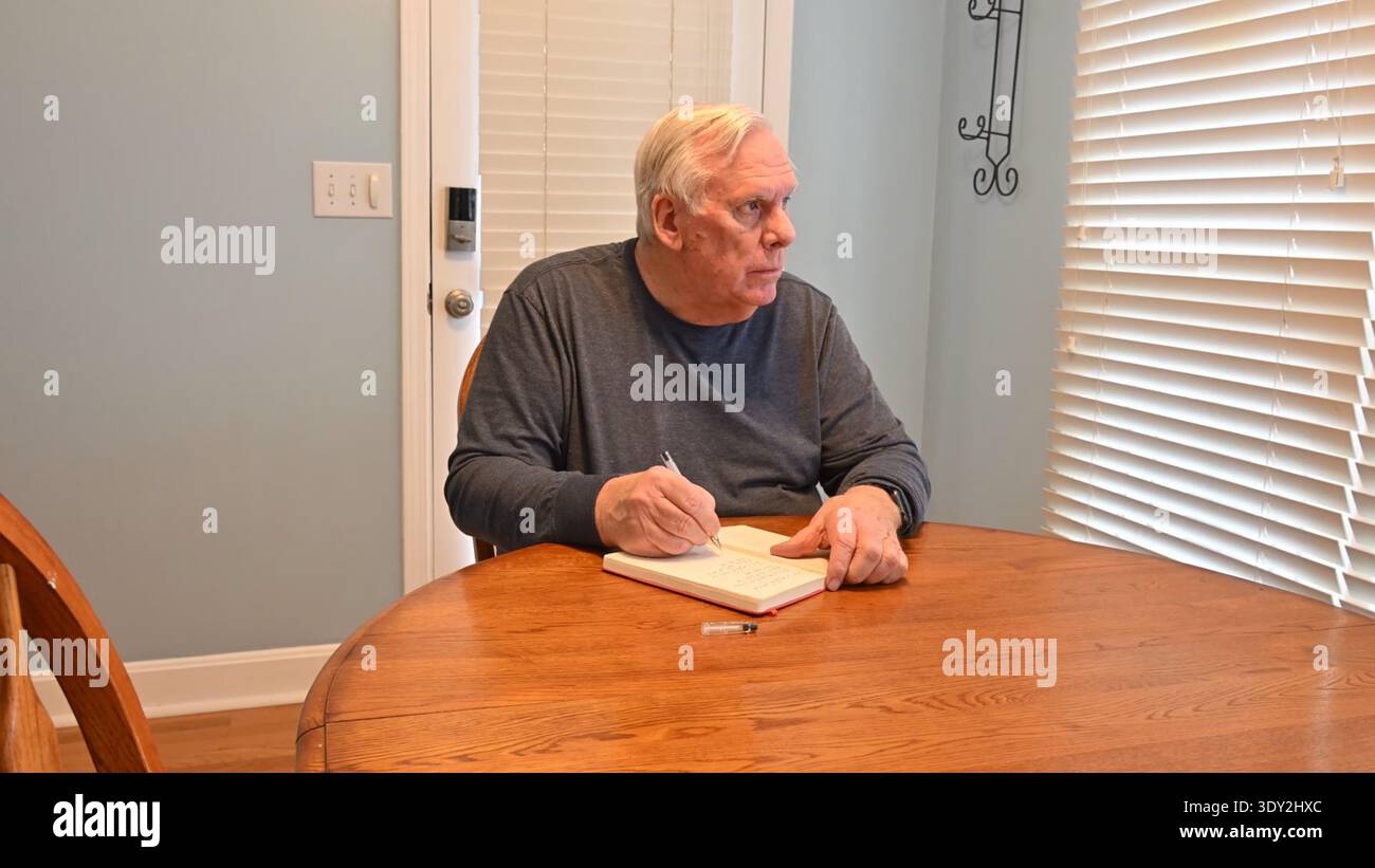 Man sits at wooden table, writing in notebook. He concentrates, eyes focused on paper. Soft light from window filters through blinds. Quiet indoor set Stock Photo