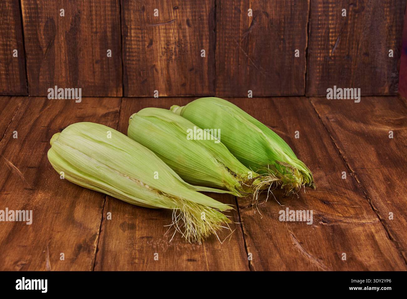 Three fresh corn cobs with green husks lying on a rustic wooden table Stock Photo