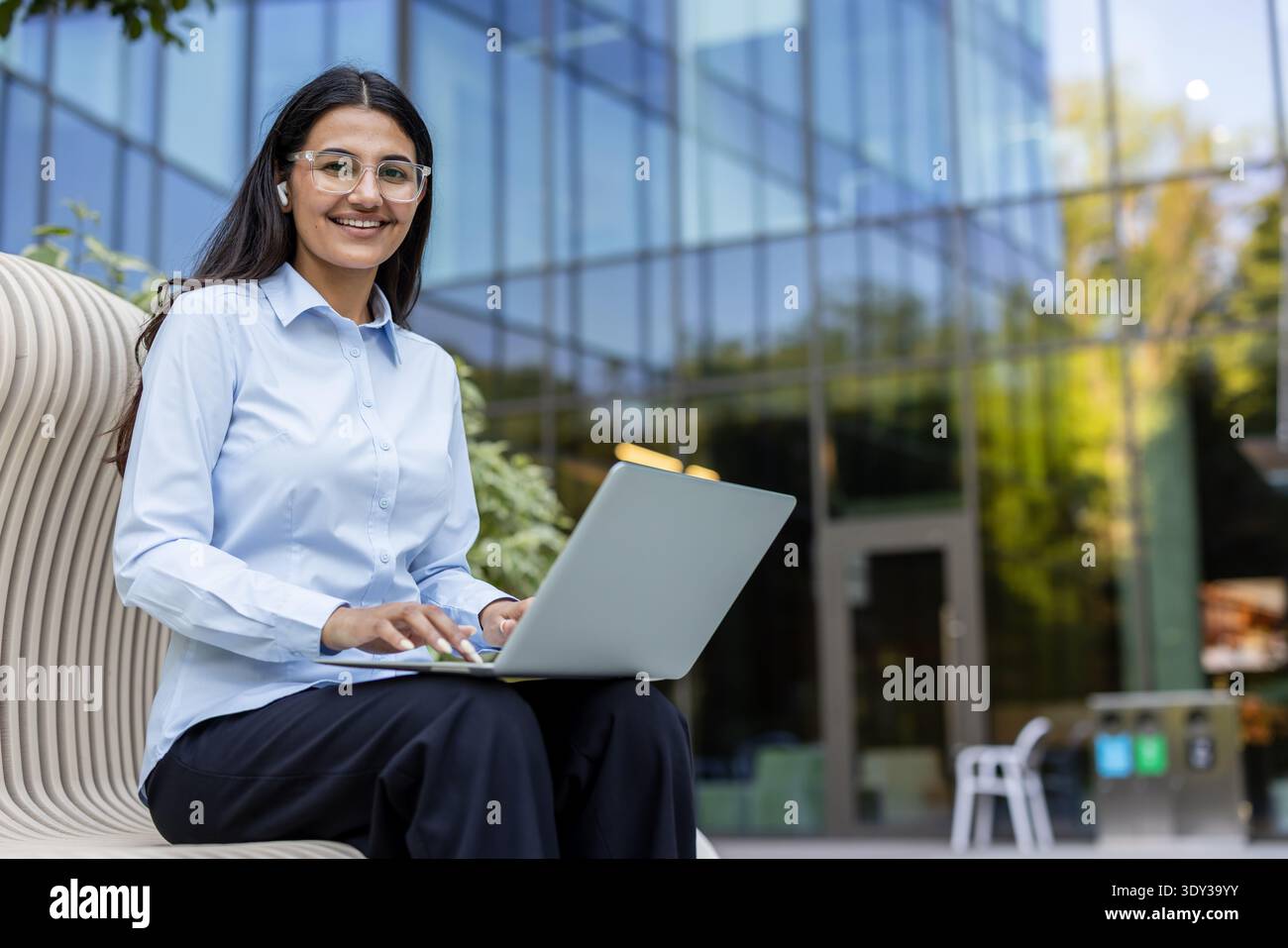 Young professional woman smiling, using a laptop and wearing earbuds while sitting on a modern bench outside a corporate office building, symbolizing remote work and technological connectivity Stock Photo