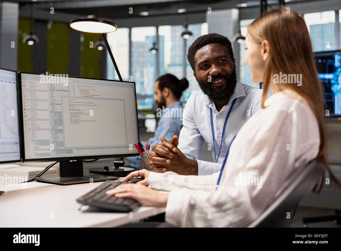 Happy senior programmer mentoring junior developer at desk in office, teaching best coding practices. Cheerful tech company lead engineer guiding intern through project tasks Stock Photo