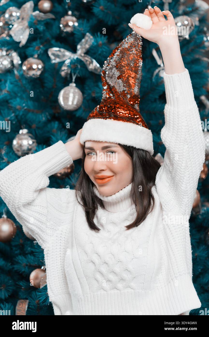 A happy young woman with blue eyes smiles while wearing a festive sequin Santa hat and a cozy white sweater. Model poses playfully in front of a decor Stock Photo