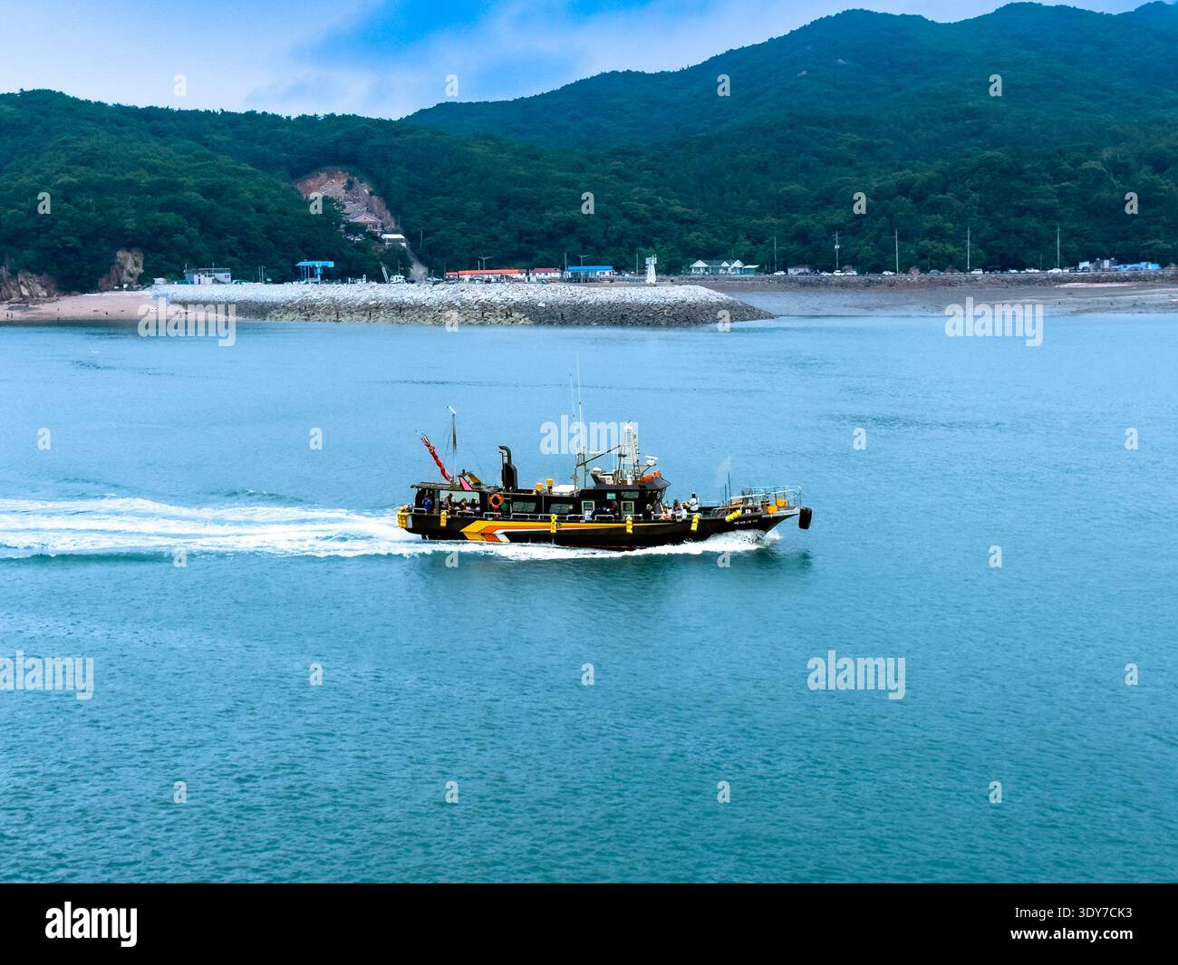 Small working boat moving across calm coastal waters with forested hills and breakwater in the background, peaceful seaside landscape. - Smartphone Captured Stock Image