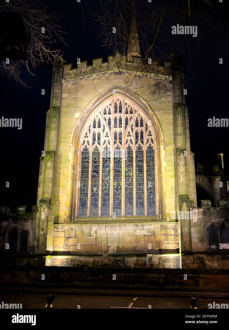 Illuminated gothic church window and facade at night with dark sky and tree branches overhead coventry uk - Smartphone Captured Stock Image