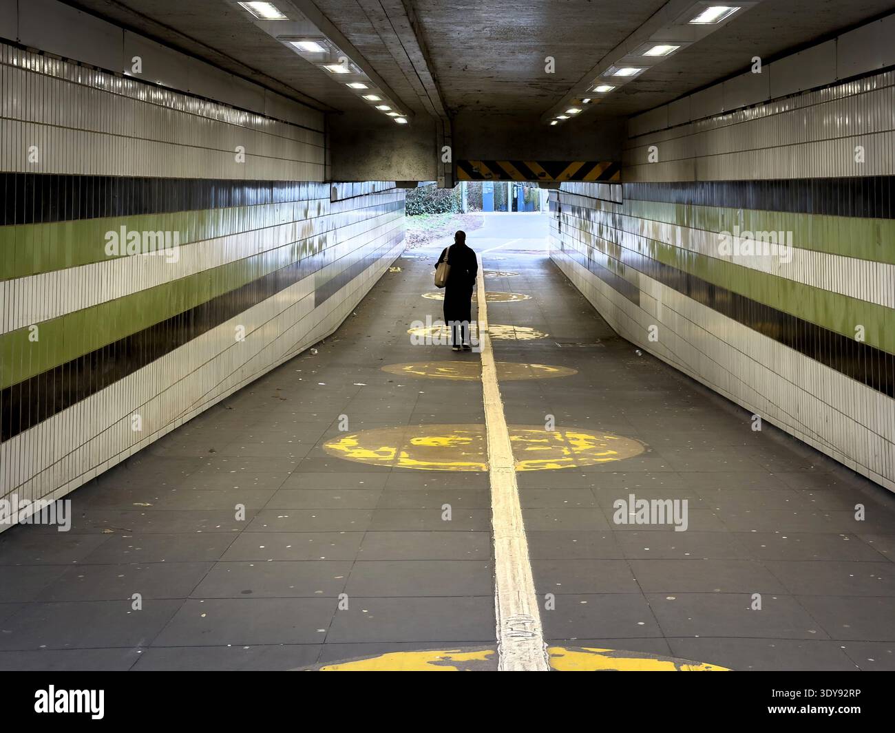 Lone pedestrian walks through a long, covered underpass with tiled walls and artificial lighting - coventry uk - Smartphone Captured Stock Image