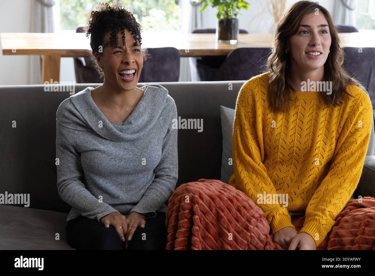 Diverse female friends laughing on gray sofa, wearing gray and mustard sweaters with rust throw Stock Photo