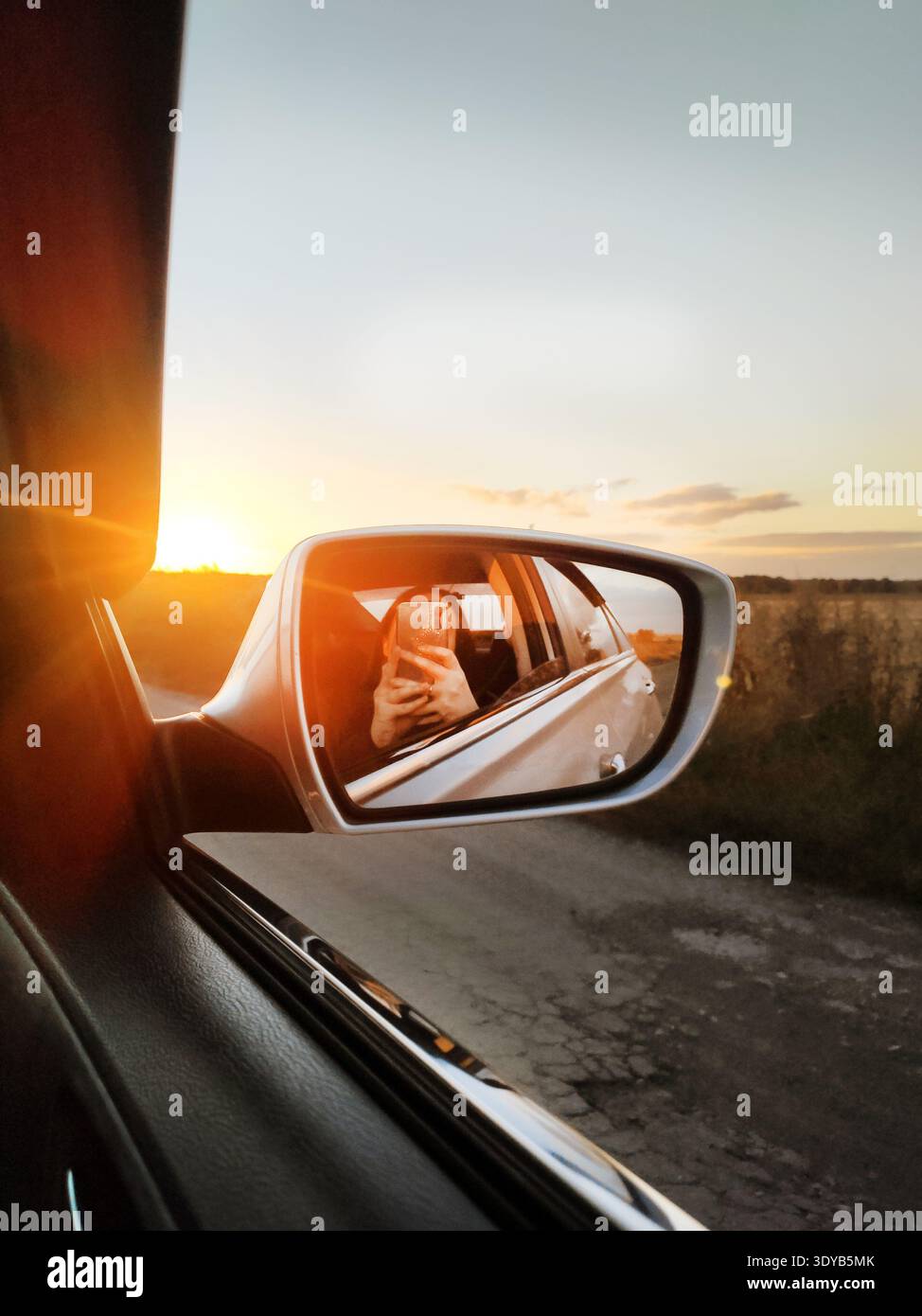 View from inside a car of the setting sun reflected in the side mirror on a country road, creating a warm atmosphere and a sun glare effect. Stock Photo