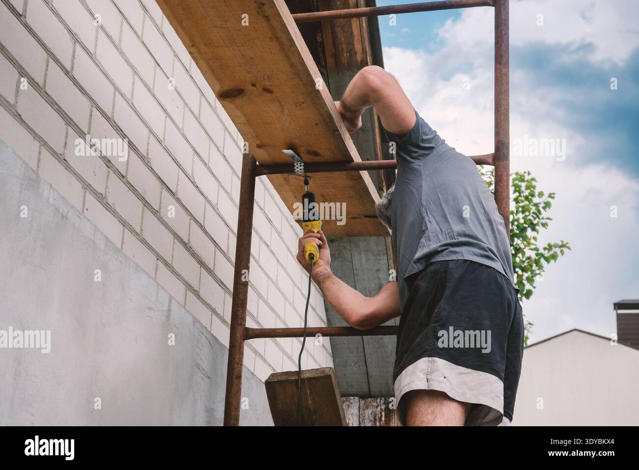 This image captures a dedicated man on scaffolding, meticulously drilling into a brick wall as part of a home renovation project. The focus highlights Stock Photo