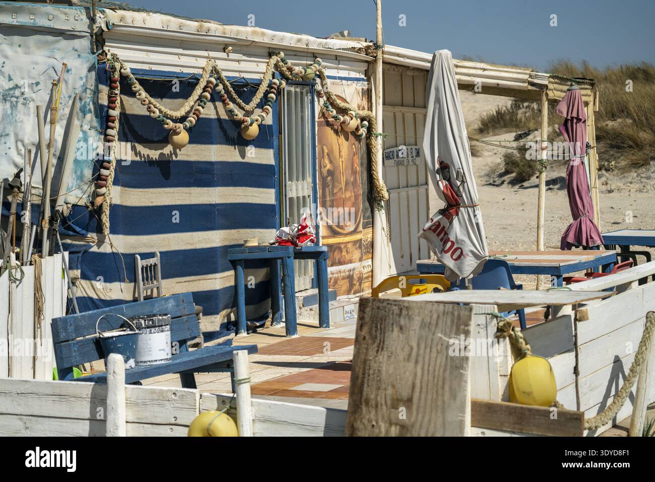 Weathered seaside structure featuring striped fabric, rope garlands, closed umbrellas, and painted religious mural under bright sunlight Stock Photo
