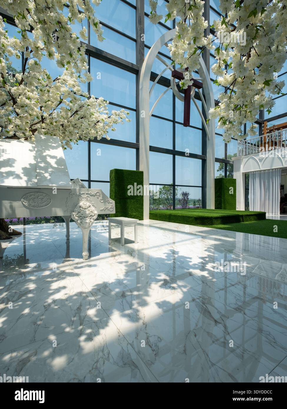 A side-angle perspective of a glass-enclosed chapel featuring a grand piano and white floral arrangements. The floor-to-ceiling glass walls offer a vi Stock Photo