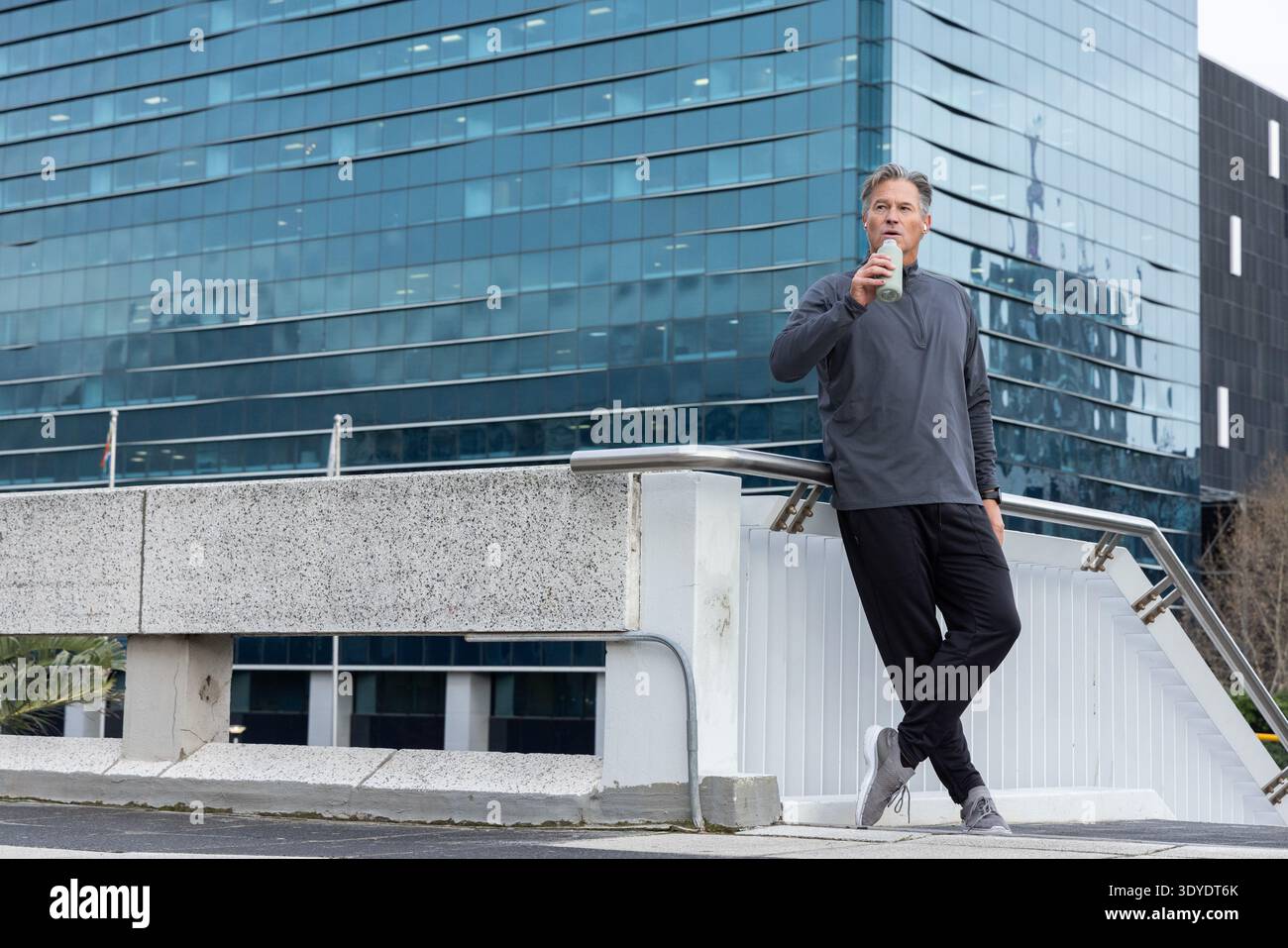 Middle-aged man in gray pullover leaning on handrail sipping bottle on overpass, copy space Stock Photo