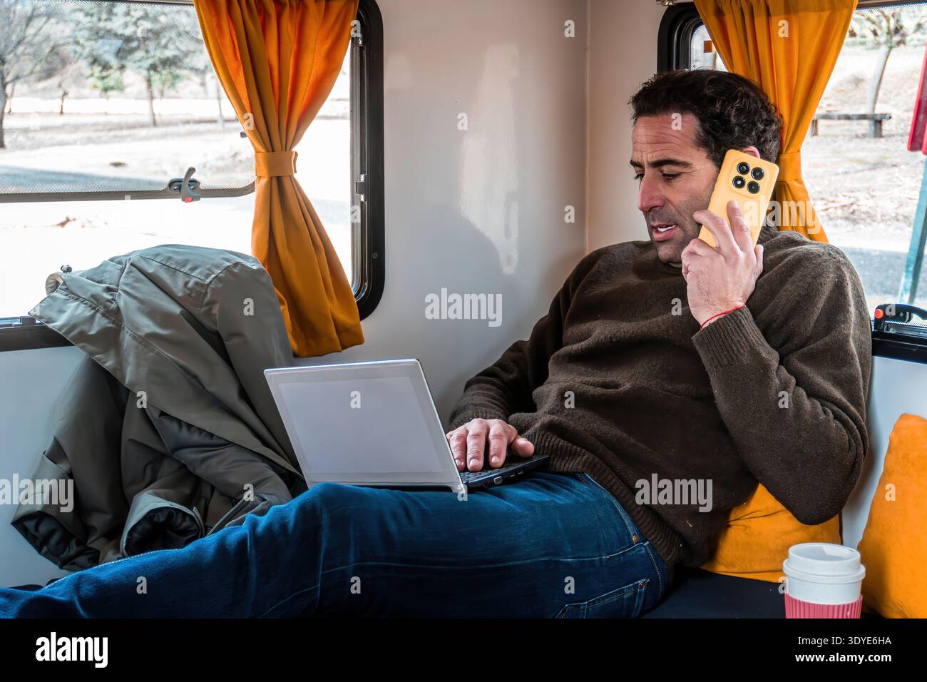 Man talking on mobile phone and using laptop inside a parked camper van. Digital nomad lifestyle, freedom and travel Stock Photo
