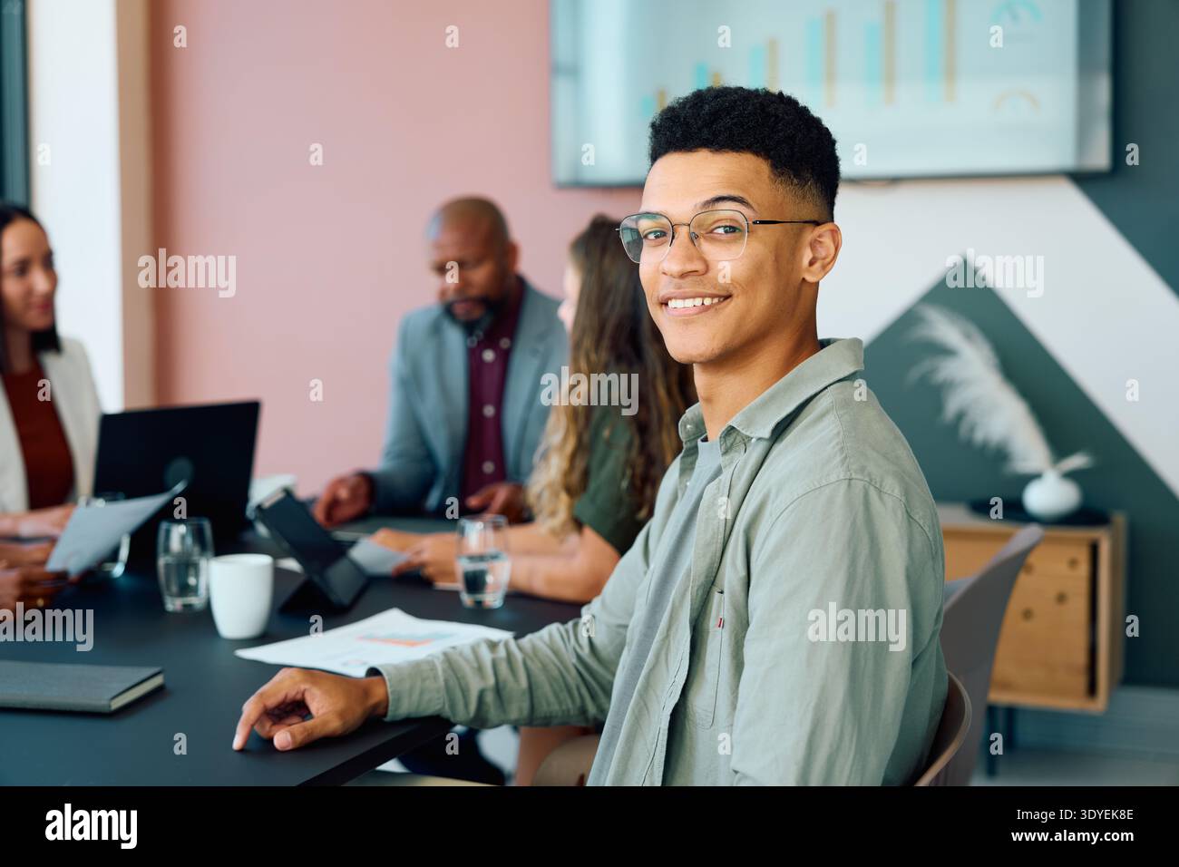 Young businessman sitting confidently and smiling at camera while team works together in boardroom at office Stock Photo